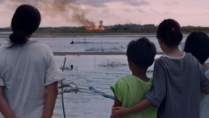 Film still from NORTE, THE END OF HISTORY: You can see a group of people from behind, looking at a fire on the other side of the river.