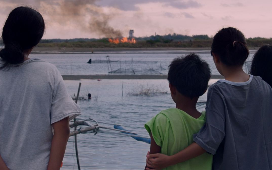 Film still from NORTE, THE END OF HISTORY: You can see a group of people from behind, looking at a fire on the other side of the river.