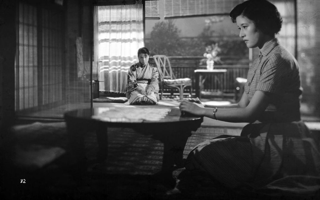 Two women are sitting in a room. One in the foreground at a low table, the other in the background.