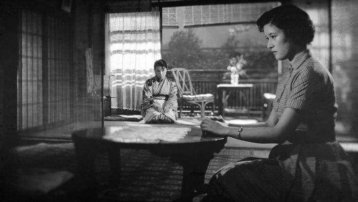 Two women are sitting in a room. One in the foreground at a low table, the other in the background.