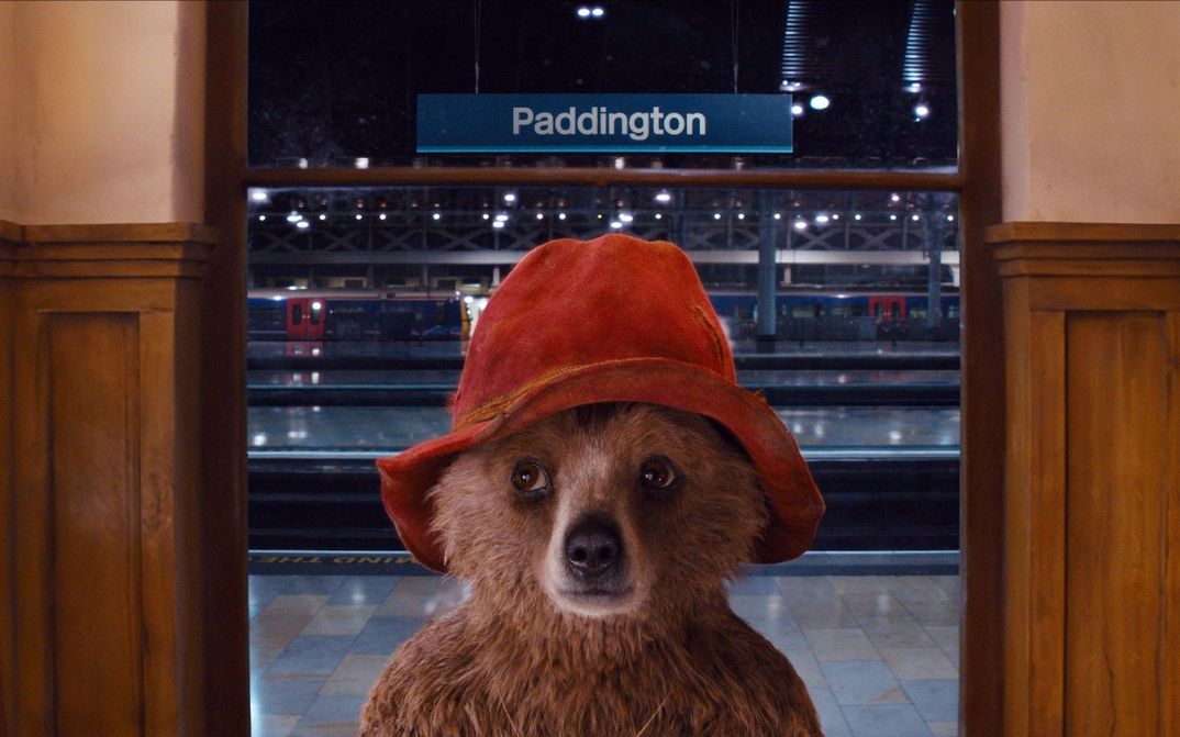 Filmstill aus PADDINGTON: Ein Teddybär mit einem roten Hut steht in einem Bahnhof. Hinter ihm sieht man die Gleise und eine Tafel mit der Aufschrift "Paddington".