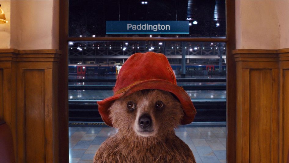 Film still from PADDINGTON: A teddy bear wearing a red hat is standing in a train station. Behind him, you can see the tracks and a sign that says “Paddington.”