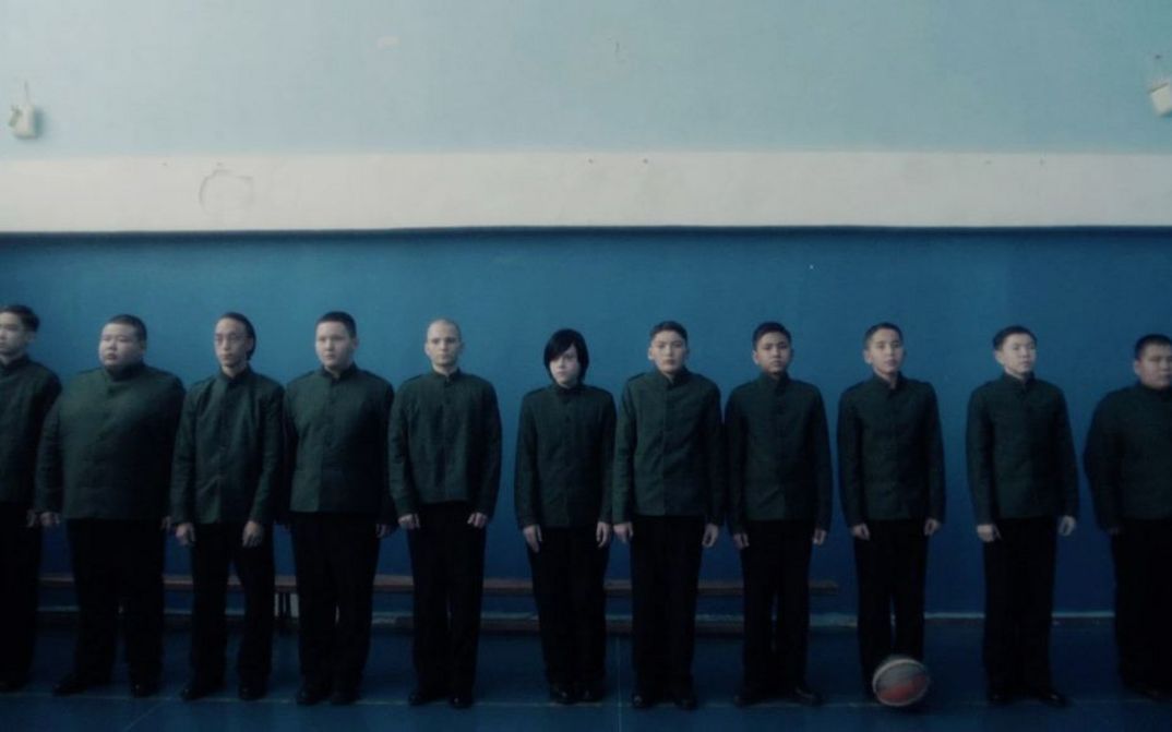 Film still from CADET by Adilkhan Yerzhanov. A group of pupils in school uniforms stand lined up in front of a wall. One of them has longer hair than the others.