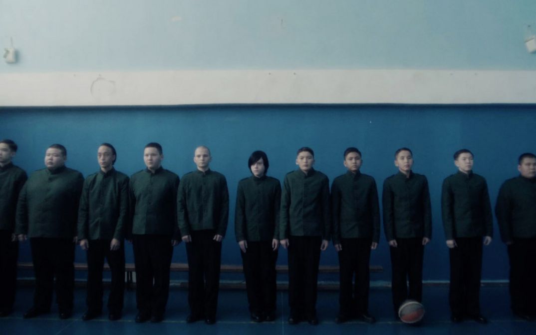 Film still from CADET by Adilkhan Yerzhanov. A group of pupils in school uniforms stand lined up in front of a wall. One of them has longer hair than the others.
