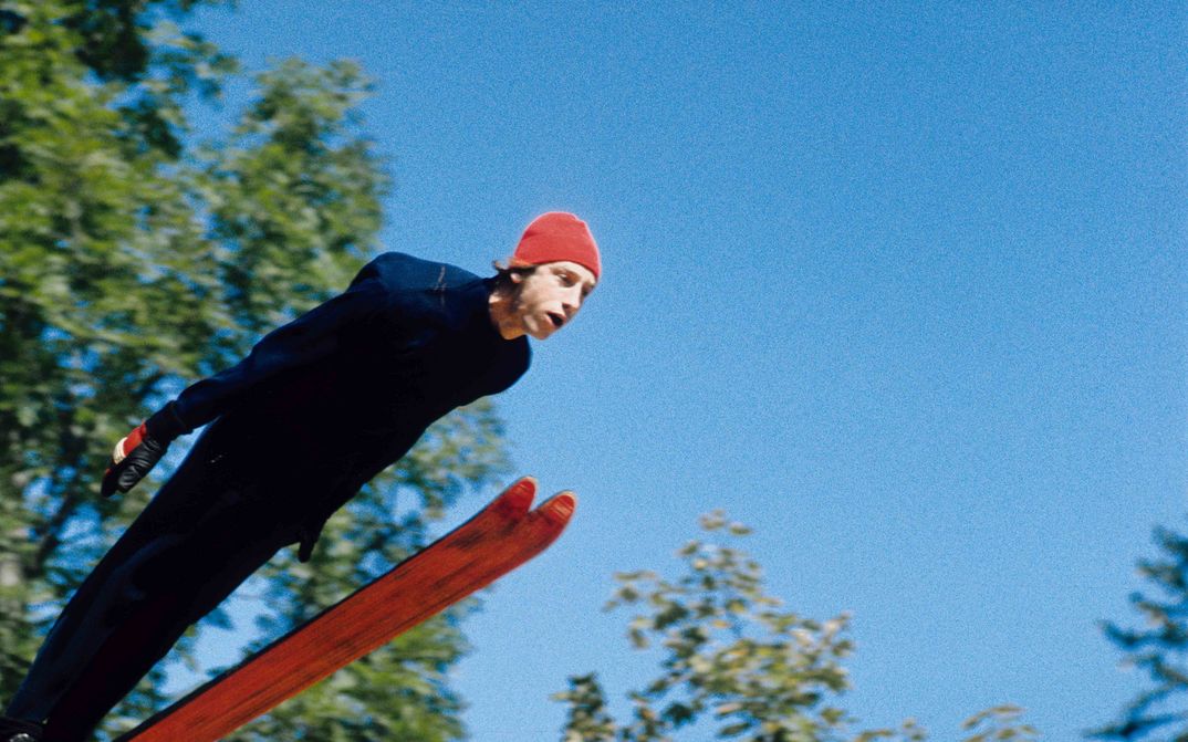 Filmstill aus DIE GROSSE EKSTASE DES BILDSCHNITZERS STEINER: Ein Skispringer mit roter Mütze, hinter ihm blauer Himmel und die grünen Blätter von Bäumen.