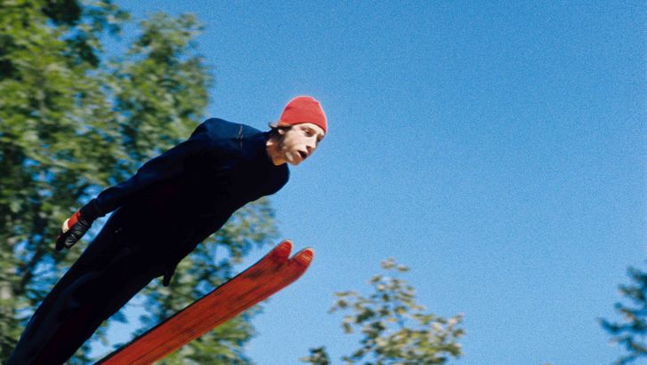 Film still from DIE GROSSE EKSTASE DES BILDSCHNITZERS STEINER: A ski jumper with red cap, behind him blue sky and the green leaves of trees.