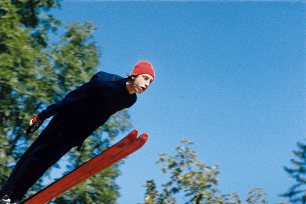 Filmstill aus DIE GROSSE EKSTASE DES BILDSCHNITZERS STEINER: Ein Skispringer mit roter Mütze, hinter ihm blauer Himmel und die grünen Blätter von Bäumen.
