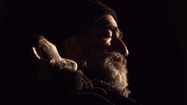 Film still from MES FANTÔMES ARMENIENS by Tamara Stepanyan. It shows the side profile of a smiling man who is slightly illuminated in the darkness. 