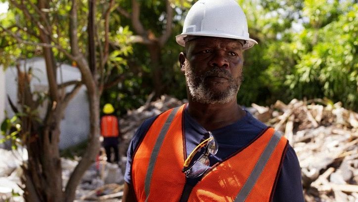 Film still from MOUNTAINS: A man wearing a safety helmet and vest stands in front of a demolished building.