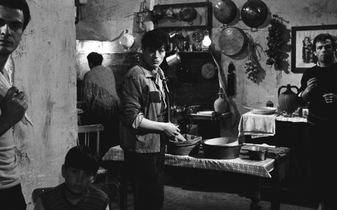 Filmstill from ROCCO E I SUOI FRATELLI: Several men and a woman in the kitchen. There are pots on the table and some kitchen utensils and decorations hanging on the wall.