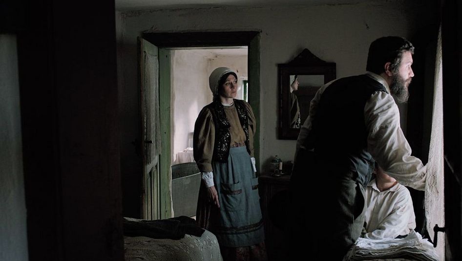 Film still from "Săptămâna Mare" by Andrei Cohn. It shows a dark room with a man and a child at the window and a woman on the left in the doorway. They are all looking out of the window. 