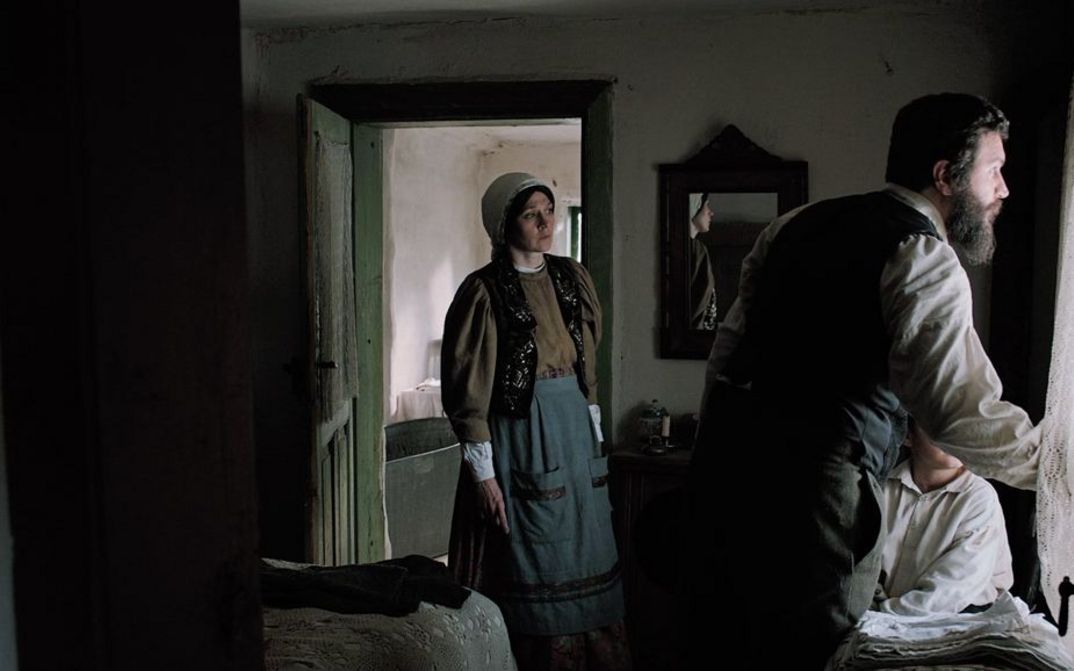 Filmstill aus "Săptămâna Mare" von Andrei Cohn. Zu sehen ist ein dunkler Raum mit einem Mann und einem Kind am Fenster und einer Frau links in der Tür. Sie alle schauen aus dem Fenster. 