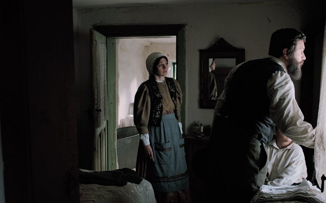 Filmstill aus "Săptămâna Mare" von Andrei Cohn. Zu sehen ist ein dunkler Raum mit einem Mann und einem Kind am Fenster und einer Frau links in der Tür. Sie alle schauen aus dem Fenster.