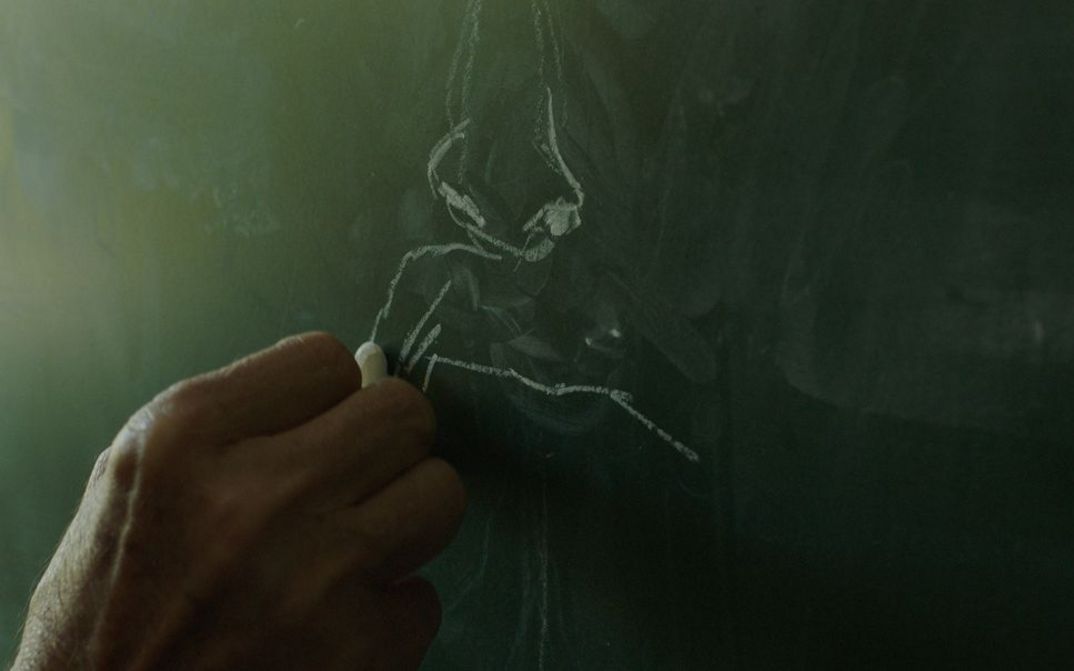 Filmstill aus dem Film „Mua besoj më shpëtoj portreti“ von Alban Muja. Eine Hand zeichnet in Nahaufnahme mit Kreide auf eine Tafel.