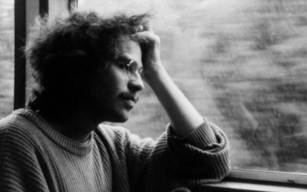 Filmstill aus IM ÜBERGANG: Ein Mann mit Brille sitzt am Fenster im Zug, stützt seinen Kopf auf der Hand ab und schaut aus dem Fenster.