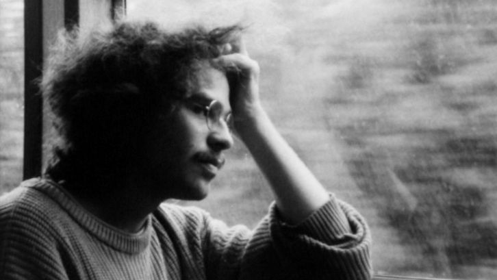 Filmstill aus IM ÜBERGANG: Ein Mann mit Brille sitzt am Fenster im Zug, stützt seinen Kopf auf der Hand ab und schaut aus dem Fenster.