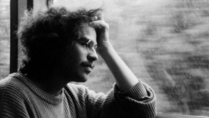 Film still from IM ÜBERGANG: A man with glasses sits at the window in the train, rests his head on his hand and looks out of the window.