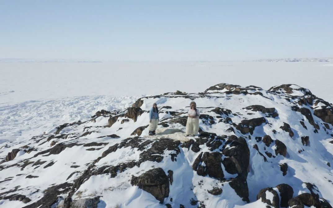 Filmstill aus „Tartupaluk (Prototype)“ von Laakkuluk Williamson Bathory. Vogelperspektive auf eine Gesteins- und Schneelandschaft. Ein großes Fell auf dem Boden, daneben zwei Personen in Fellhosen und Fellmützen die uns anschauen.
