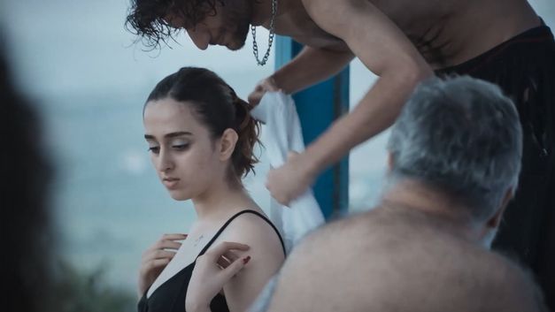 Mehrere Personen sitzen eng beieinander, ein Mann legt einer Frau ein Tuch um die Schultern. Im Hintergrund ist eine Wasserfläche.