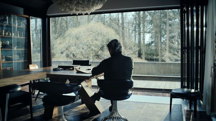 Filmstill aus LA MORT VIENDRA: In einer Wohnung mit einer riesigen Fensterfront, hinter der man einen Wald sieht, sitzt ein Mann an einem Tisch und blickt nach draußen.