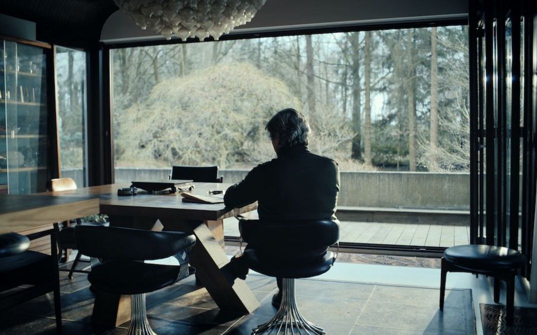Film still from LA MORT VIENDRA: In an apartment with a huge window that overlooks a forest, a man is sitting at a table, looking outside.