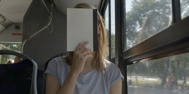 Filmstill aus „We Haven’t Lost Our Way“ von Anka und Wilhelm Sasnal. Eine Frau sitzt im Bus und hält ein Notizbuch vor ihr Gesicht. 