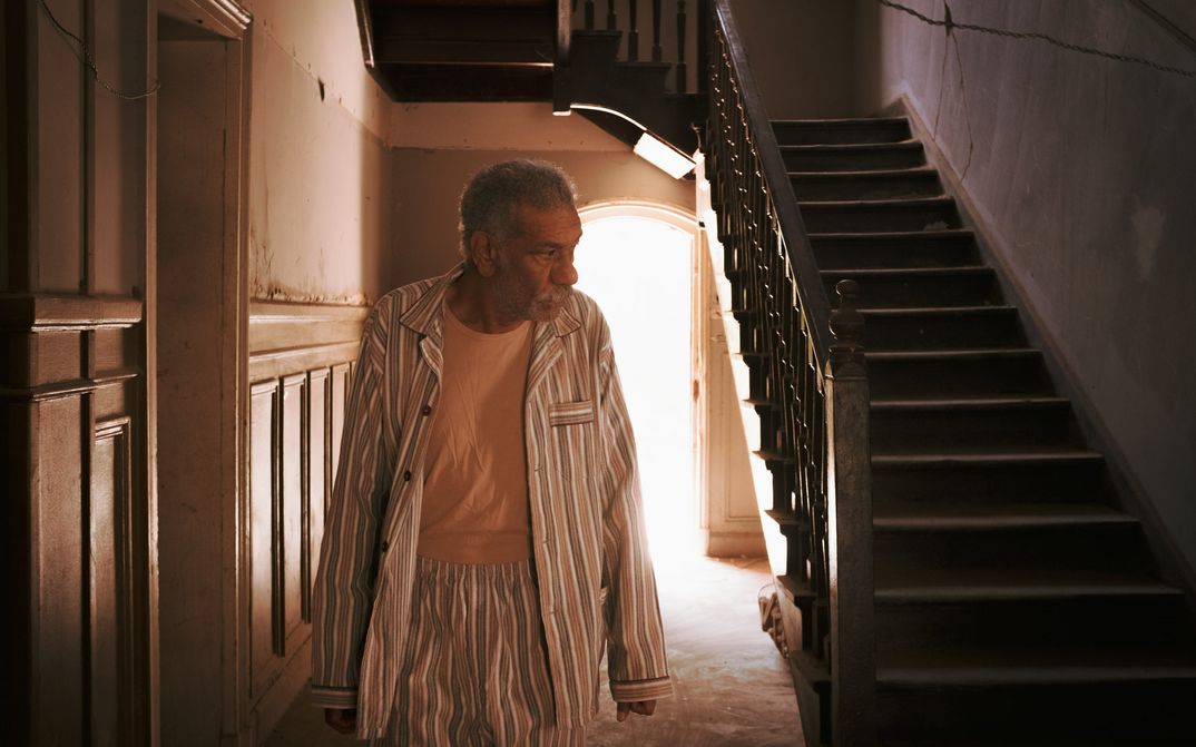 Film still from 19B: An elderly man stands in his pyjamas in a stairwell.