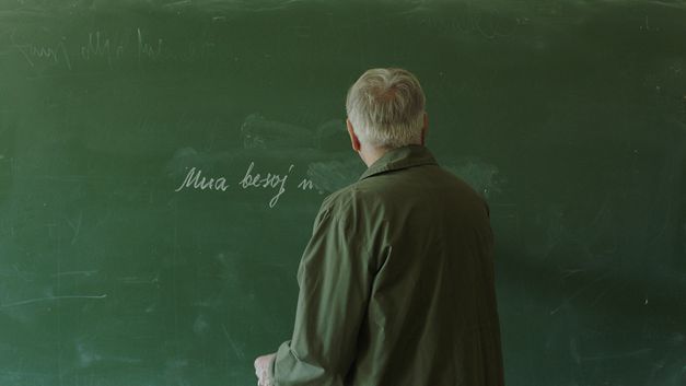 Film still from Alban Muja’s film “Mua besoj më shpëtoj portreti”. A middle-aged man, seen from behind, writes with chalk on a blackboard.