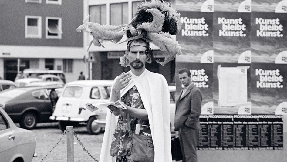 Jack Smith steht mit einem weißen Umhang und einem aufwendigen Kopfschmuck mit Federn auf der Straße. Im Hintergrund sind Häuser, Autos, Menschen und Plakate mit der Aufschrift Kunst bleibt Kunst. zu sehen.