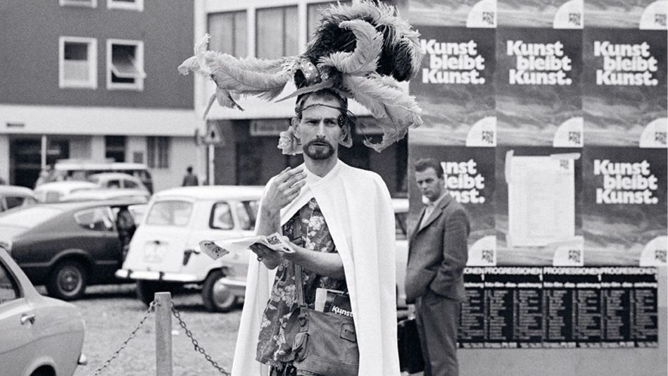  Jack Smith stands on the street wearing a white cape and an elaborate headdress adorned with feathers. In the background, you can see houses, cars, people, and posters bearing the slogan “Art remains art.”