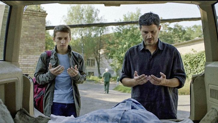Film still from HOMEWARD: A father and his son stand in front of the open boot of their car and say a prayer.