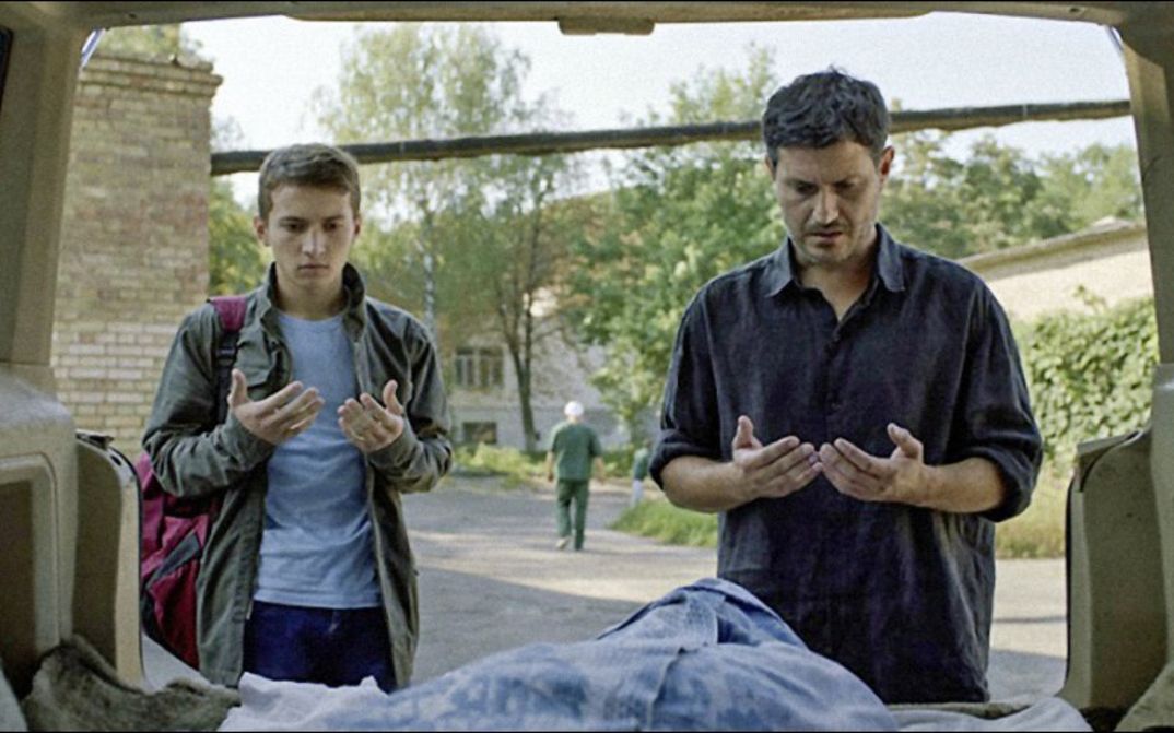 Film still from HOMEWARD: A father and his son stand in front of the open boot of their car and say a prayer.