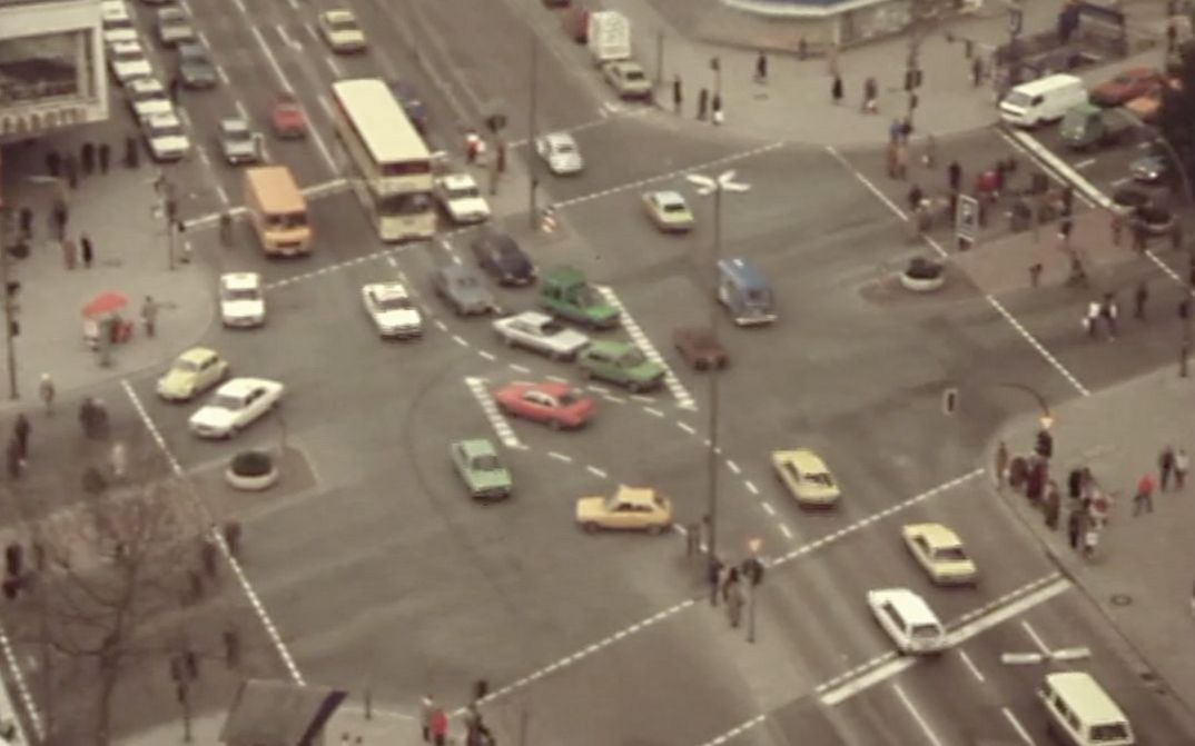 Still from the film "Merry Christmas Deutschland oder Vorlesung zur Geschichtstheorie II" by Raoul Peck. Aerial view of an intersection in an metropolitan area: cars, busses, pedestrians.