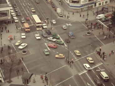 Still from the film "Merry Christmas Deutschland oder Vorlesung zur Geschichtstheorie II" by Raoul Peck. Aerial view of an intersection in an metropolitan area: cars, busses, pedestrians. 