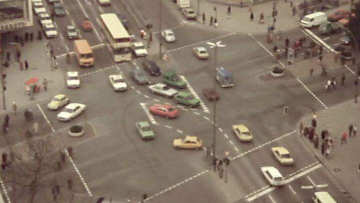 Still from the film "Merry Christmas Deutschland oder Vorlesung zur Geschichtstheorie II" by Raoul Peck. Aerial view of an intersection in an metropolitan area: cars, busses, pedestrians. 