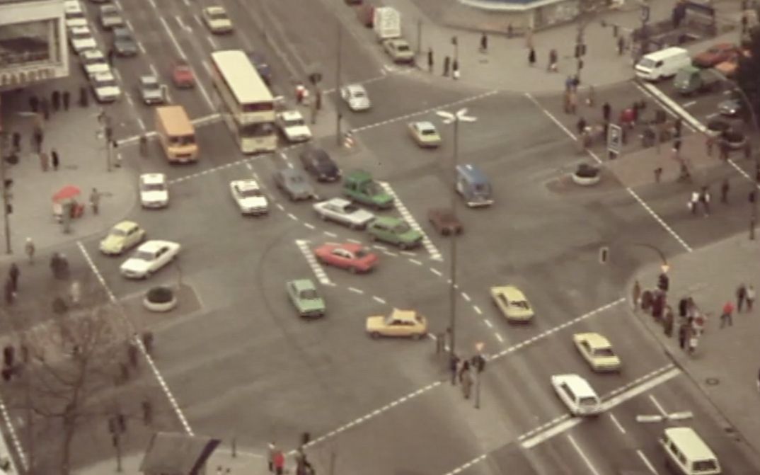 Still from the film "Merry Christmas Deutschland oder Vorlesung zur Geschichtstheorie II" by Raoul Peck. Aerial view of an intersection in an metropolitan area: cars, busses, pedestrians. 