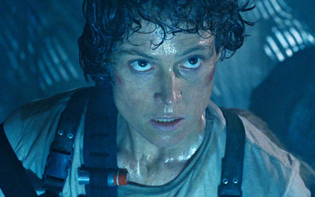Film still from ALIENS: A woman, covered in sweat and armed with a cartridge belt, in a bluish-dark room.
