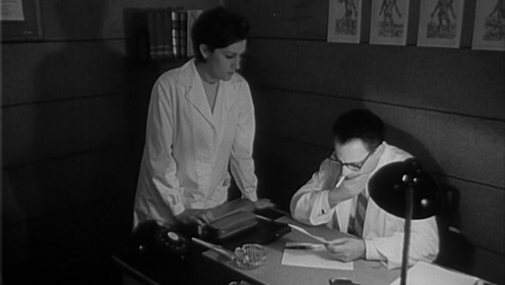 Filmstill aus DOMINGO À TARDE: Ein Arzt sitzt rauchend und lesend am Schreibtisch, neben ihm steht eine Frau.
