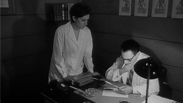Film still from DOMINGO À TARDE: A doctor is sitting at his desk smoking and reading, a woman is standing next to him.