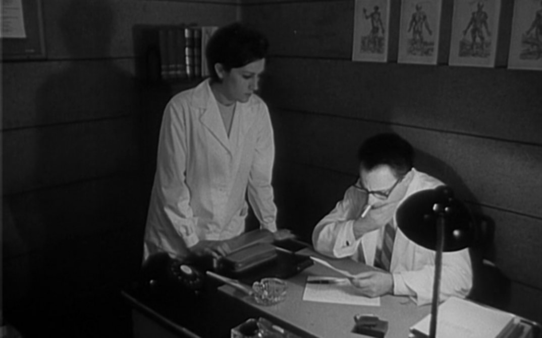 Film still from DOMINGO À TARDE: A doctor is sitting at his desk smoking and reading, a woman is standing next to him.