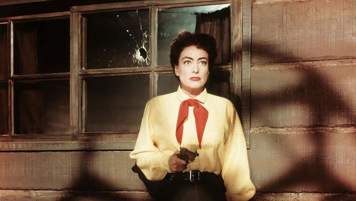 Filmstill aus JOHNNY GUITAR: Eine Frau mit gelbem Hemd und rotem Halstuch hält eine Pistole in der Hand, schaut erschrocken und steht vor einem Holzhaus und einem Fenster, welches ein Einschussloch hat.