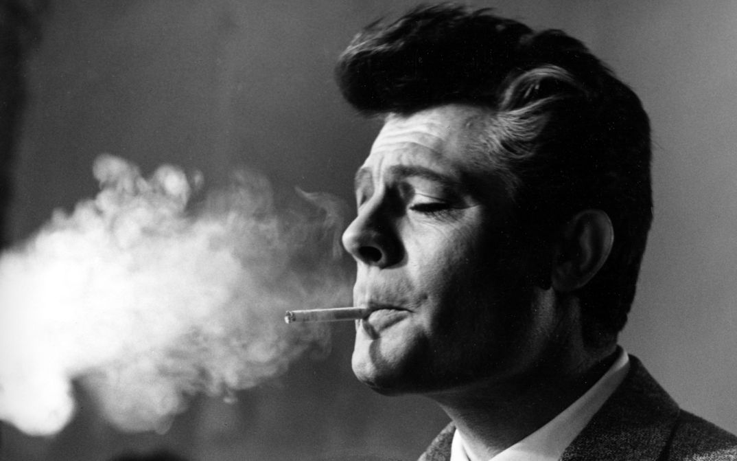 Black and white photograph of Marcello Mastroianni smoking a cigarette. The smoke fills the left part of the photo.