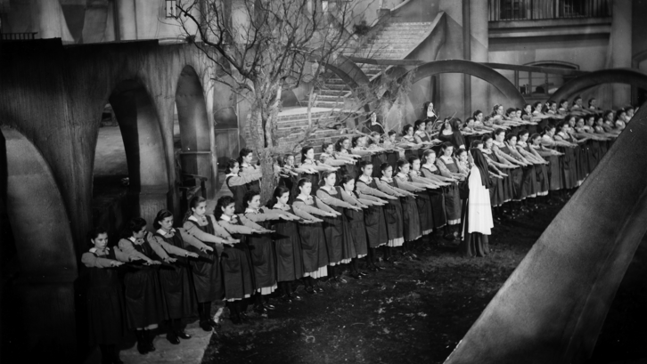 Film still from MUCHACHAS DE UNIFORME: Schoolgirls in uniform are lined up in an expressionist setting. A nun inspects their outstretched hands.