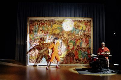 Film still from "Shahid" by Narges Kalhor. It shows a man in front of a printed screen illuminated by a spotlight. To his left is a man sitting at the drums.