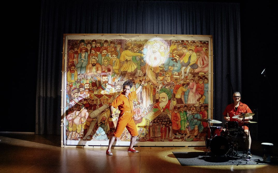 Filmstill aus "Shahid" von Narges Kalhor. Zu sehen ist ein Mann vor einem gedruckten Leinwand, das von einem Scheinwerfer angestrahlt wird. Zu seiner Linken sitzt ein Mann an einem Schlagzeug.