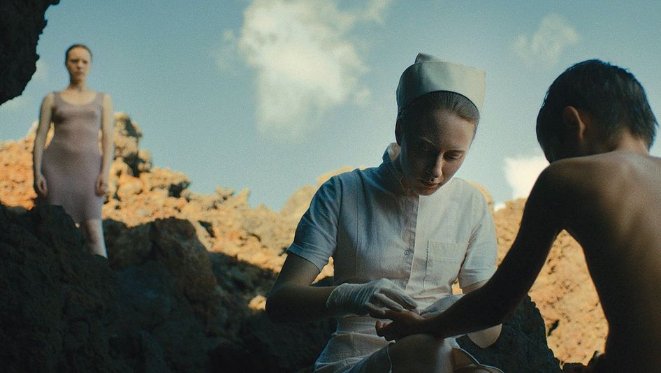 Filmstill aus ÉVOLUTION: Eine Krankenschwester in weißer Haube verarztet einen Jungen auf einer Klippe. Eine junge Frau im Hintergrund schaut zu.