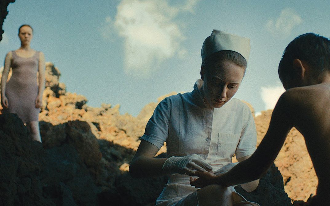Film still from ÉVOLUTION: A nurse treats a boy on a cliff. A young woman watches in the background.