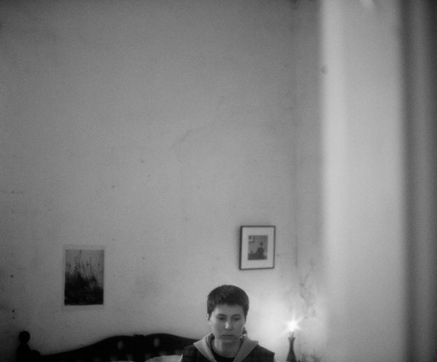 Film still from BATIM by Veronica Nicole Tetelbaum. The black and white image shows a room with high ceilings and two pictures on the wall. At the bottom of the picture are the head of a person and part of a bed.