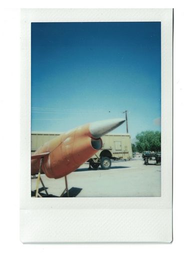 Polaroid einer oragnefarbenen Rakete vor einem beigen Lastwagen und blauem Himmel.