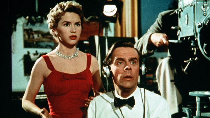 Filmstill aus SIMON AND LAURA: Eine Frau in einem eleganten roten Kleid und ein Mann in einem weißen Hemd mit Fliege stehen neben einer großen Filmkamera und blicken auf das Gefilmte.